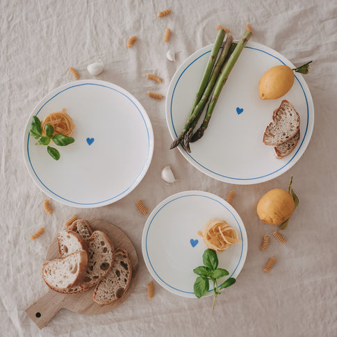 Mehrere Porzellanteller mit hellblauem Rand und Herzmotiv in verschiedenen Größen, arrangiert mit Brot, Spargel und Zitronen.