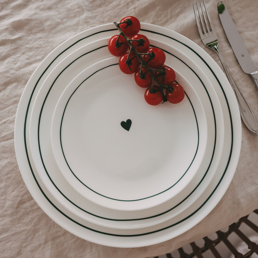Stapel weißer Porzellanteller mit grünem Rand und Herzdetail, dekoriert mit Cherrytomaten und Besteck.
