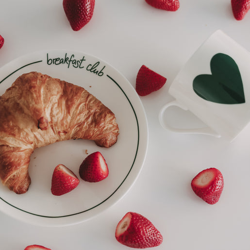 Frühstücksteller „breakfast club“ mit Croissant und Erdbeeren, daneben Tasse mit grünem Herzmotiv auf hellem Hintergrund
