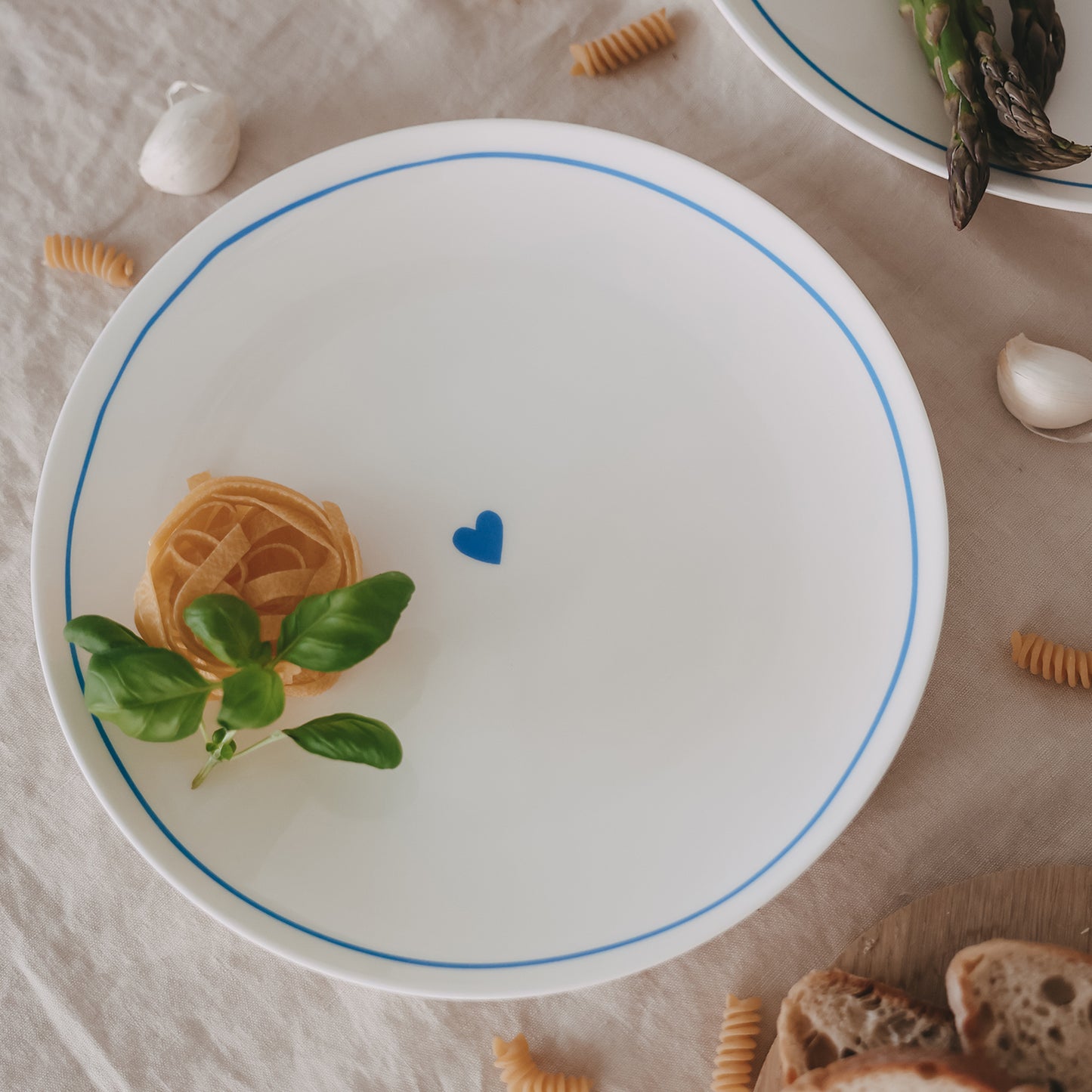Großer Porzellanteller mit hellblauem Rand und Herzmotiv, dekoriert mit Pasta und Basilikum auf Leinentuch.