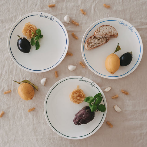 Mehrere „dinner club“-Teller mit buntem Rand auf Leinentisch, dekoriert mit Pasta, Brot, Zitrone, Aubergine und verstreuten Nudeln und Knoblauch.
