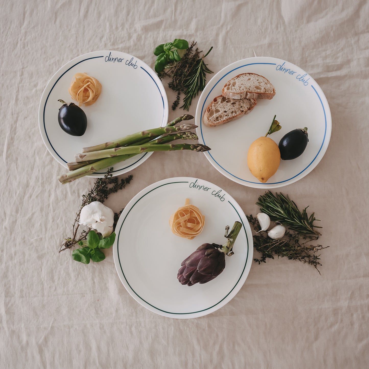 3 „dinner club“-Teller mit buntem Rand, angerichtet mit Spargel, Brot, Zitrone und Aubergine, umgeben von Kräutern auf Leinentisch.
