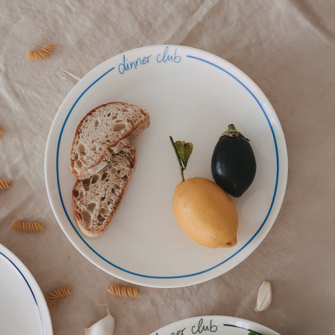 Weißer „dinner club“-Teller mit hellblauem Rand und Schrift, belegt mit Brot, Zitrone und Aubergine, auf Leinentisch mit verstreuten Nudeln und Knoblauch.