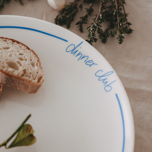 Detailaufnahme eines „dinner club“-Tellers mit hellblauer Schrift und Rand, mit Brot, Zitrone und Aubergine, umgeben von frischen Kräutern.