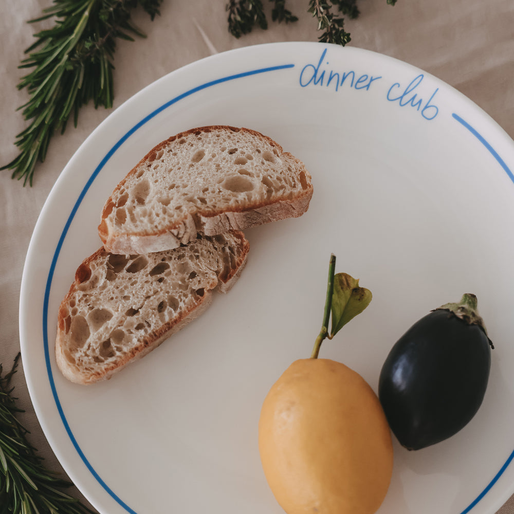 Nahaufnahme eines weißen „dinner club“-Tellers mit hellblauem Rand, angerichtet mit Brot, Zitrone und Aubergine, dekoriert mit Rosmarin und Thymian.