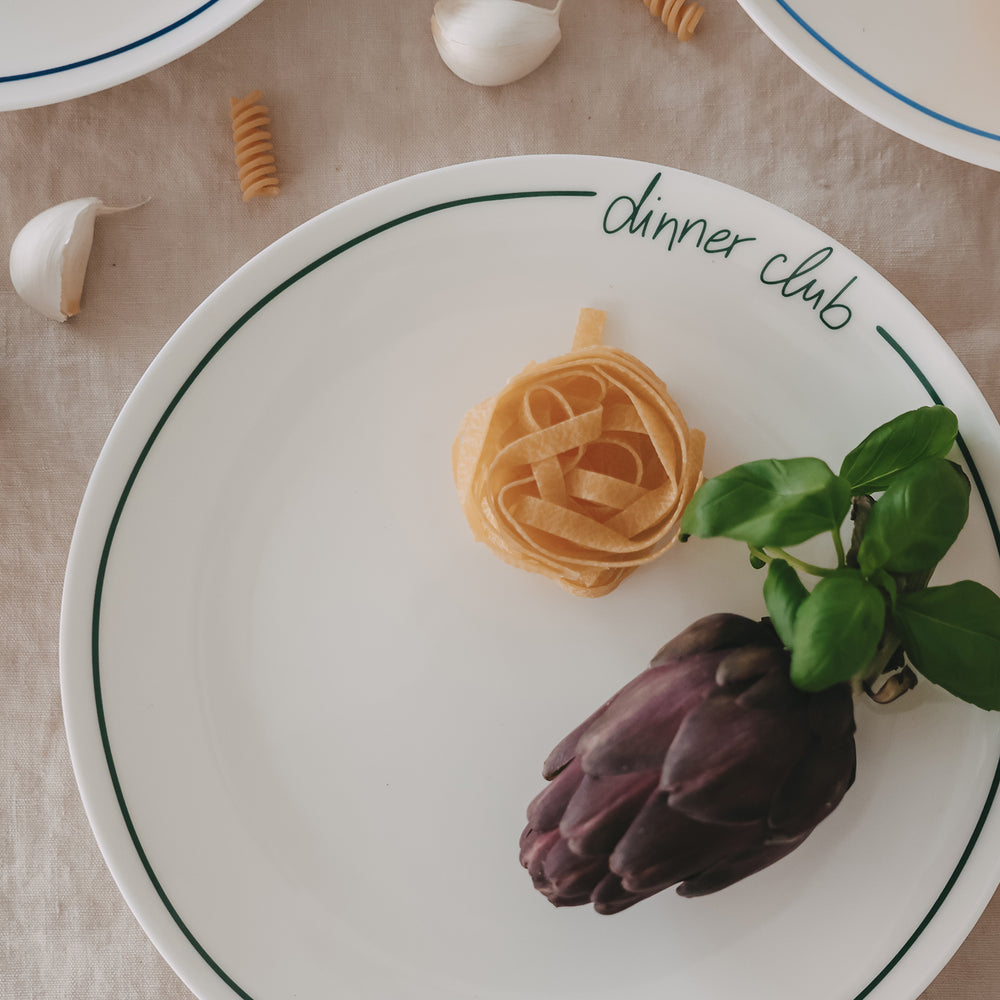 Weißer „dinner club“-Teller mit grünem Rand und Schrift, angerichtet mit Pasta-Nest, Aubergine und frischem Basilikum, auf hellem Leinentisch mit Knoblauch und Nudeln.