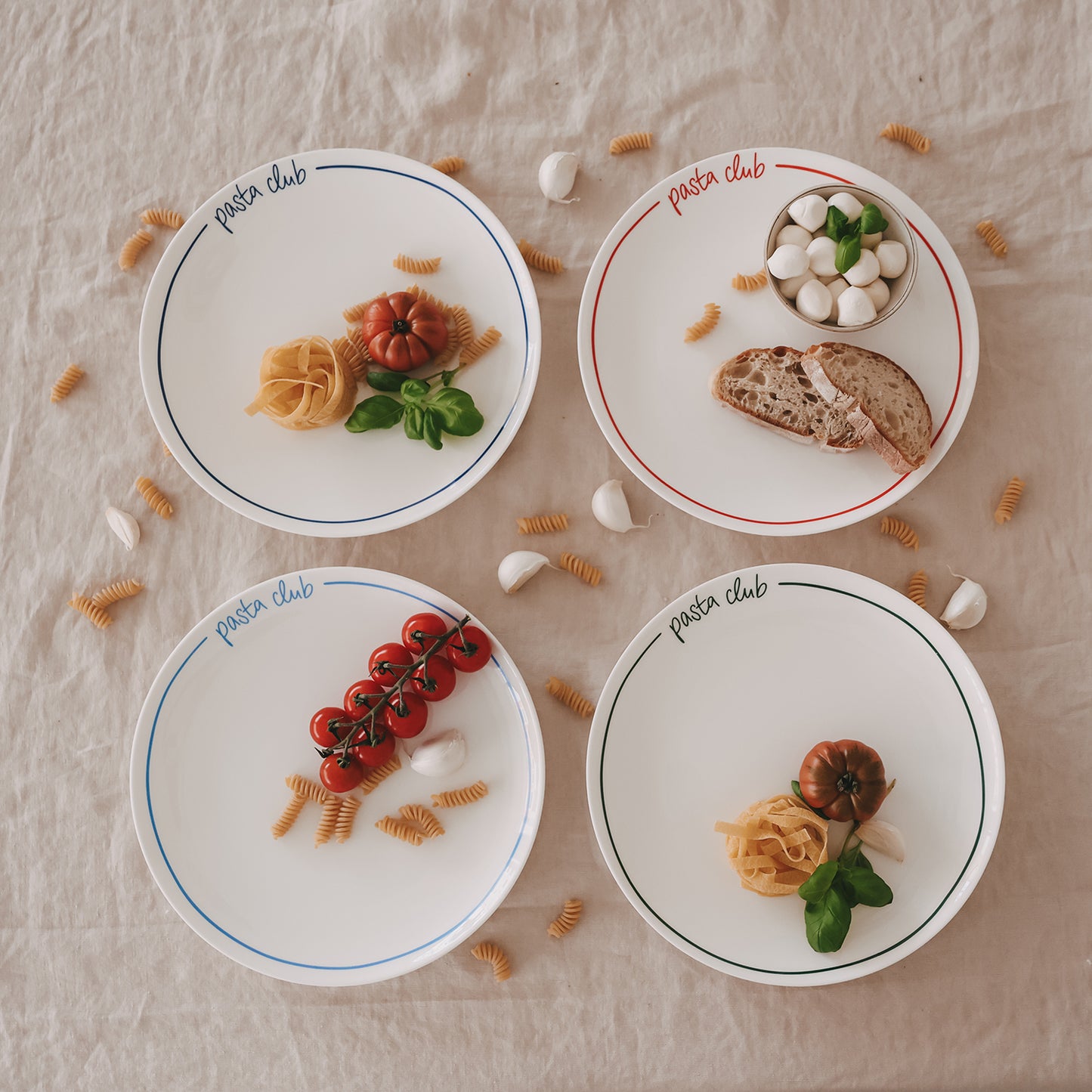 4 „pasta club“ Speiseteller in blau, hellblau, grün und rot, angerichtet mit Pasta, Brot und Mozzarella, umgeben von Knoblauch und trockenen Nudeln auf Leinentisch.