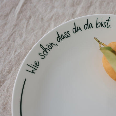 Detailansicht eines weißen Platztellers mit grüner Schrift „Wie schön, dass du da bist“ mit Zitrone, auf hellem Leinentisch.