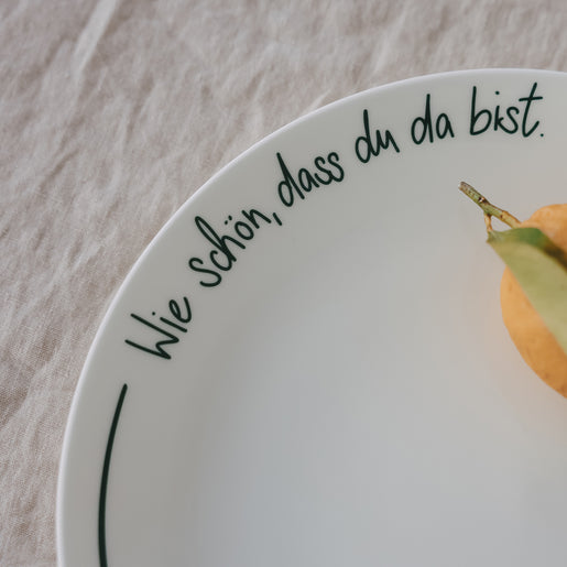 Detailansicht eines weißen Platztellers mit grüner Schrift „Wie schön, dass du da bist“ mit Zitrone, auf hellem Leinentisch.
