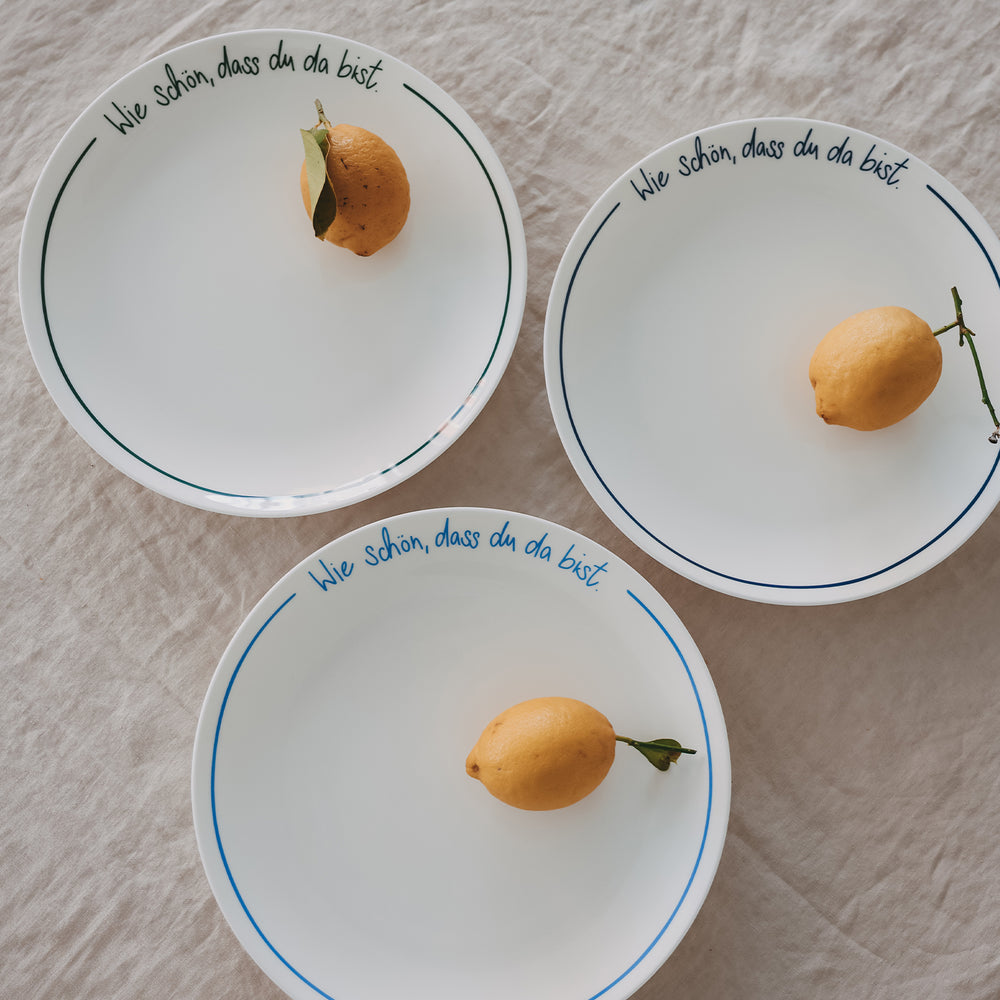 Drei Platzteller „Wie schön, dass du da bist“ in dunkelblau, grün und hellblau, jeweils mit Zitrone auf hellem Leinentisch arrangiert.