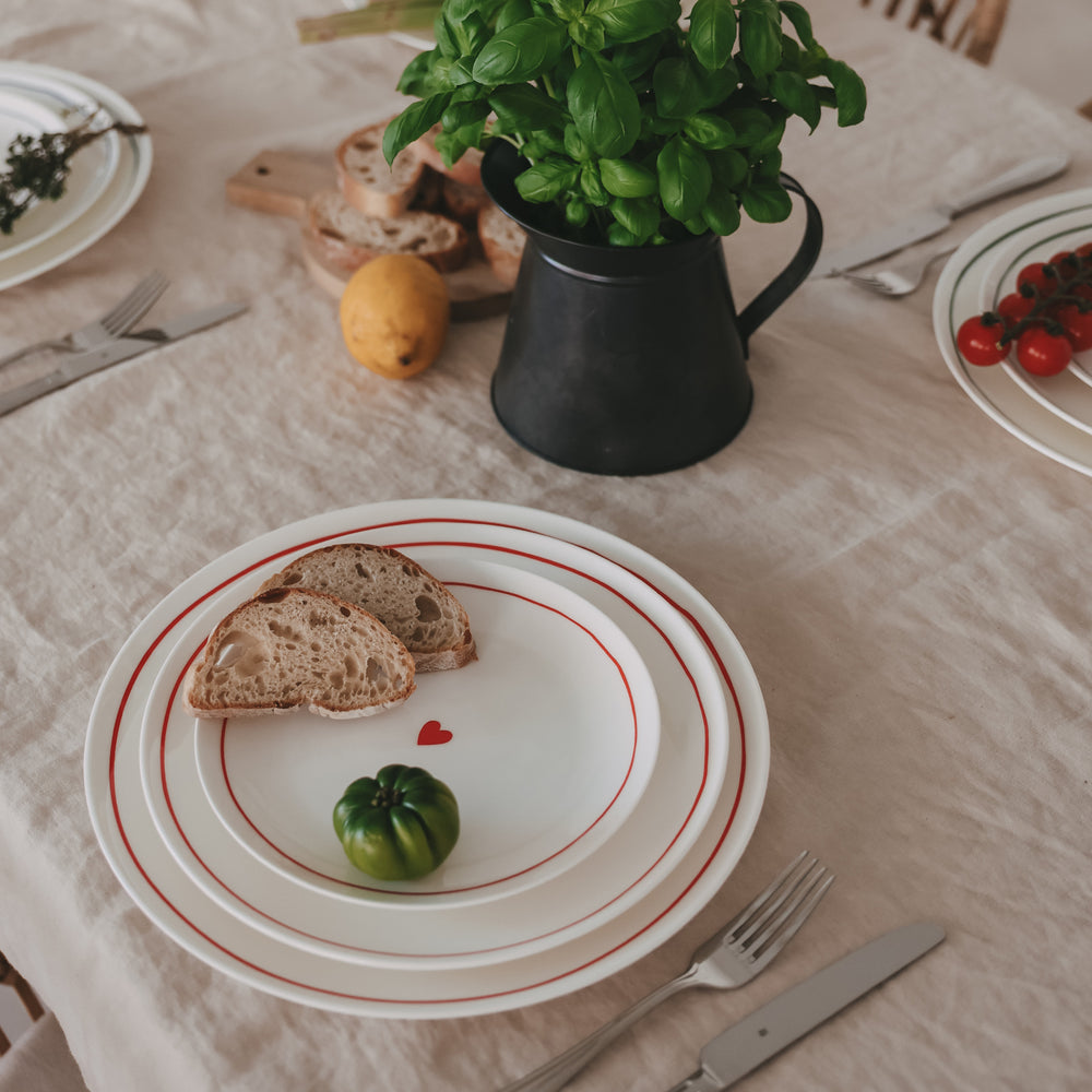 Gedeckter Tisch mit weißen Tellern mit rotem Rand und Herzdetail, dazu Brot, Basilikum in Metallkanne und frische Zutaten.