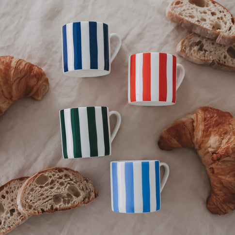 Vier kleine, gestreifte Porzellan-Tassen (dunkelblau, rot, grün, hellblau) liegen auf einem Leinentuch, umgeben von Croissants und aufgeschnittenem Brot.
