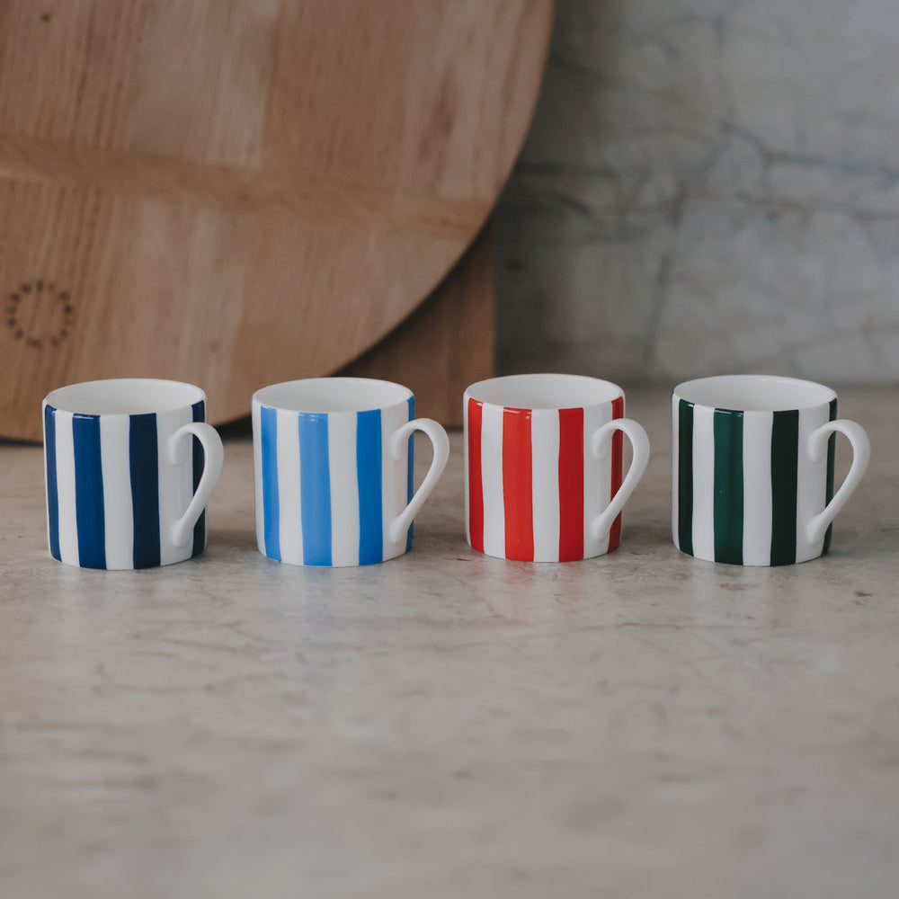 Vier kleine Tassen in einer Reihe – dunkelblau, hellblau, rot und grün – auf einer hellen Steinfläche mit Holzbrett im Hintergrund.