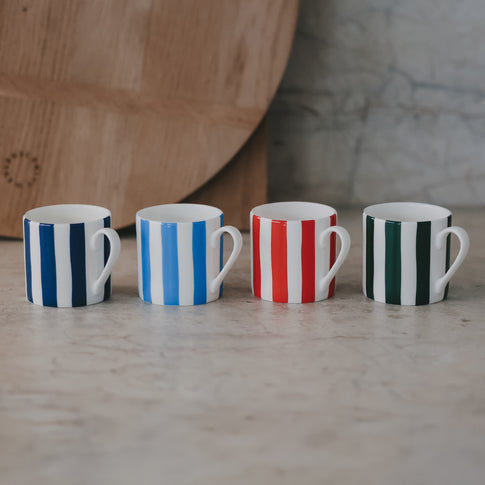 Vier kleine Tassen in einer Reihe – dunkelblau, hellblau, rot und grün – auf einer hellen Steinfläche mit Holzbrett im Hintergrund.