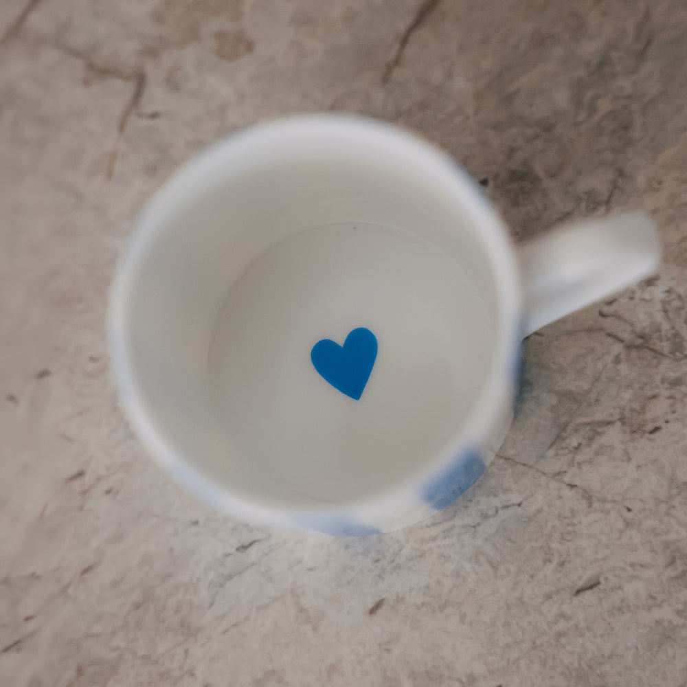 Blick in eine kleine Porzellan-Tasse mit weißem Inneren und einem kleinen blauen Herz am Boden, auf heller Steinfläche.