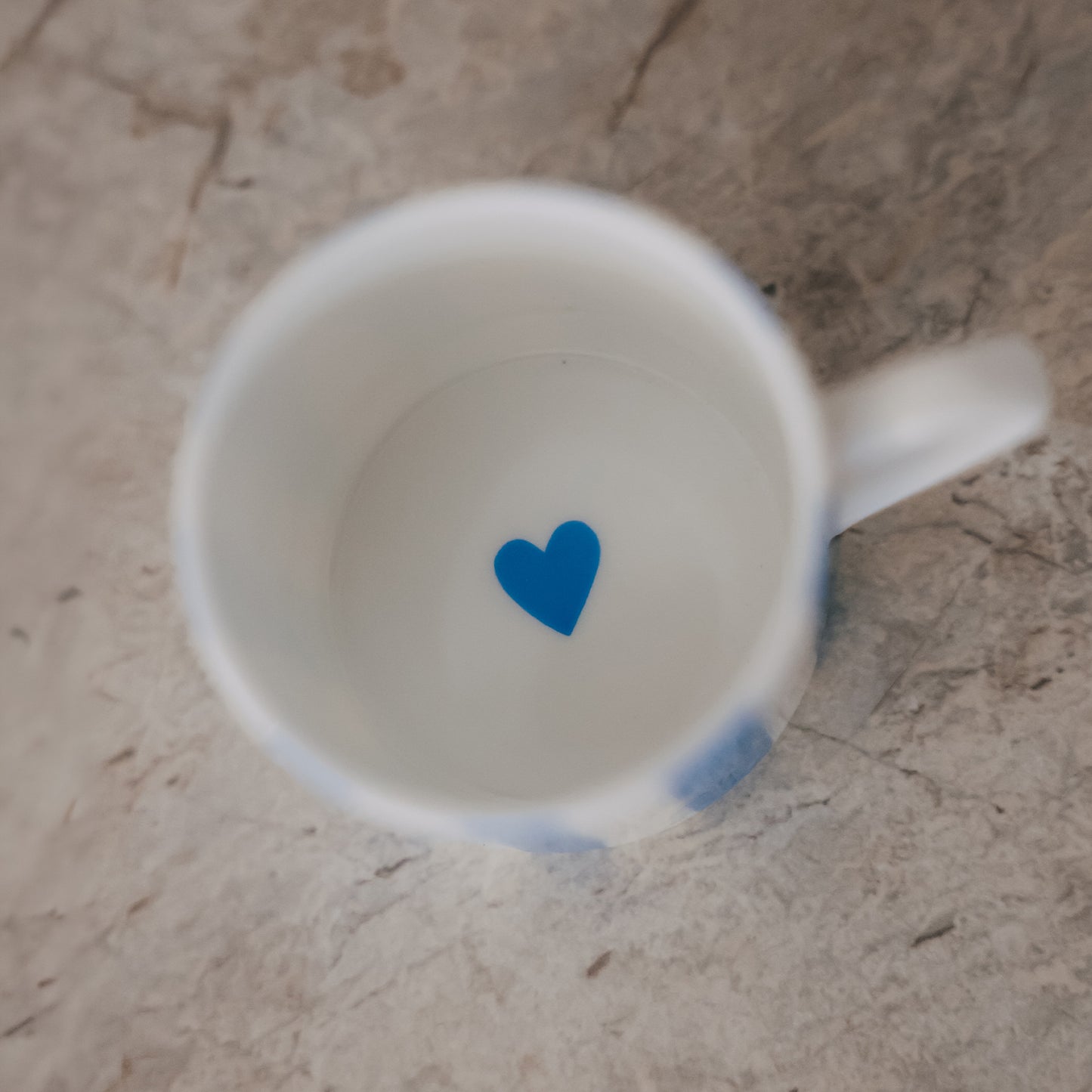 Blick in eine kleine Porzellan-Tasse mit weißem Inneren und einem kleinen blauen Herz am Boden, auf heller Steinfläche.