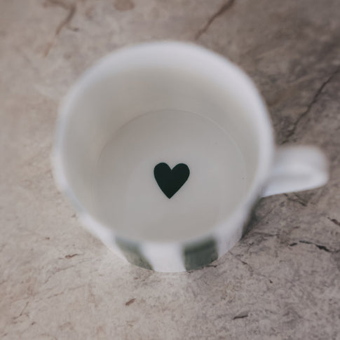 Blick in die Tasse mit weißem Inneren und kleinem grünem Herz am Boden, auf heller Steinoberfläche.