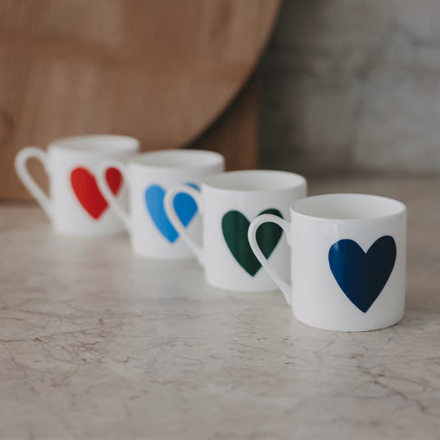 Vier kleine Tassen in einer Reihe, jeweils mit großem Herz in Rot, Hellblau, Grün und Dunkelblau, auf heller Steinfläche, weicher Hintergrund.
