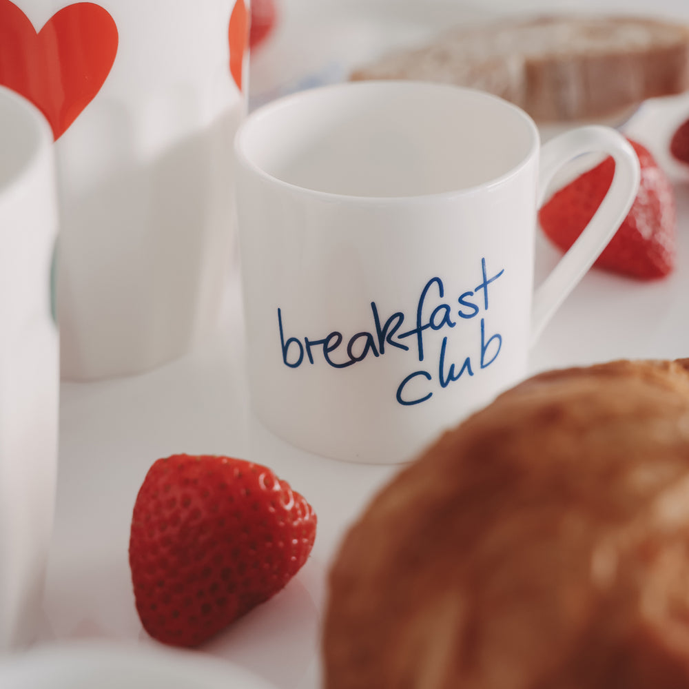 Kleine Tasse mit Schriftzug „breakfast Club“ in Blau, umgeben von Erdbeeren und Gebäck auf weißem Tisch.