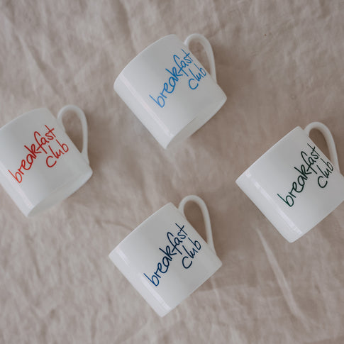 4 kleine Tassen mit „breakfast Club“-Schrift in verschiedenen Farben (rot, hellblau, dunkelblau und grün) locker auf Stoff drapiert, Draufsicht.