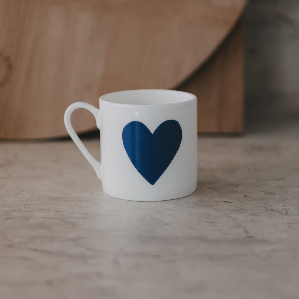 Einzelne kleine Tasse mit großem dunkelblauen Herz frontal auf heller Fläche, ruhiger Hintergrund.