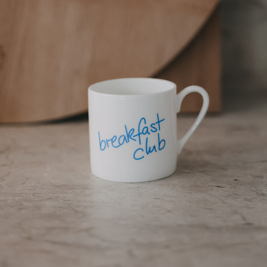 Kleine Tasse mit Schriftzug „breakfast club“ in Hellblau, seitlich fotografiert vor ruhigem Hintergrund.