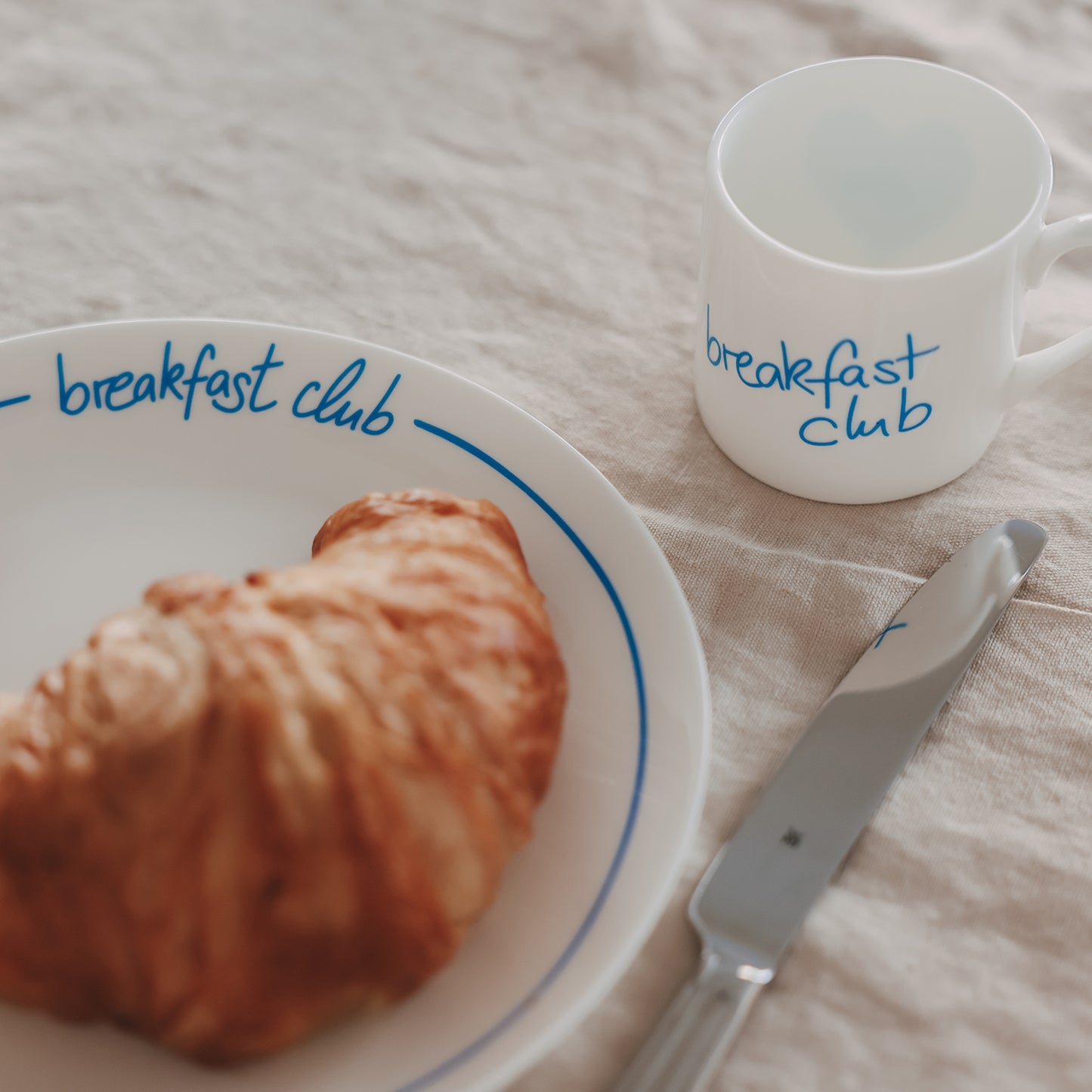 Frühstücksszene mit Croissant und Teller „breakfast club“, daneben die passende kleine Tasse mit hellblauer Schrift.