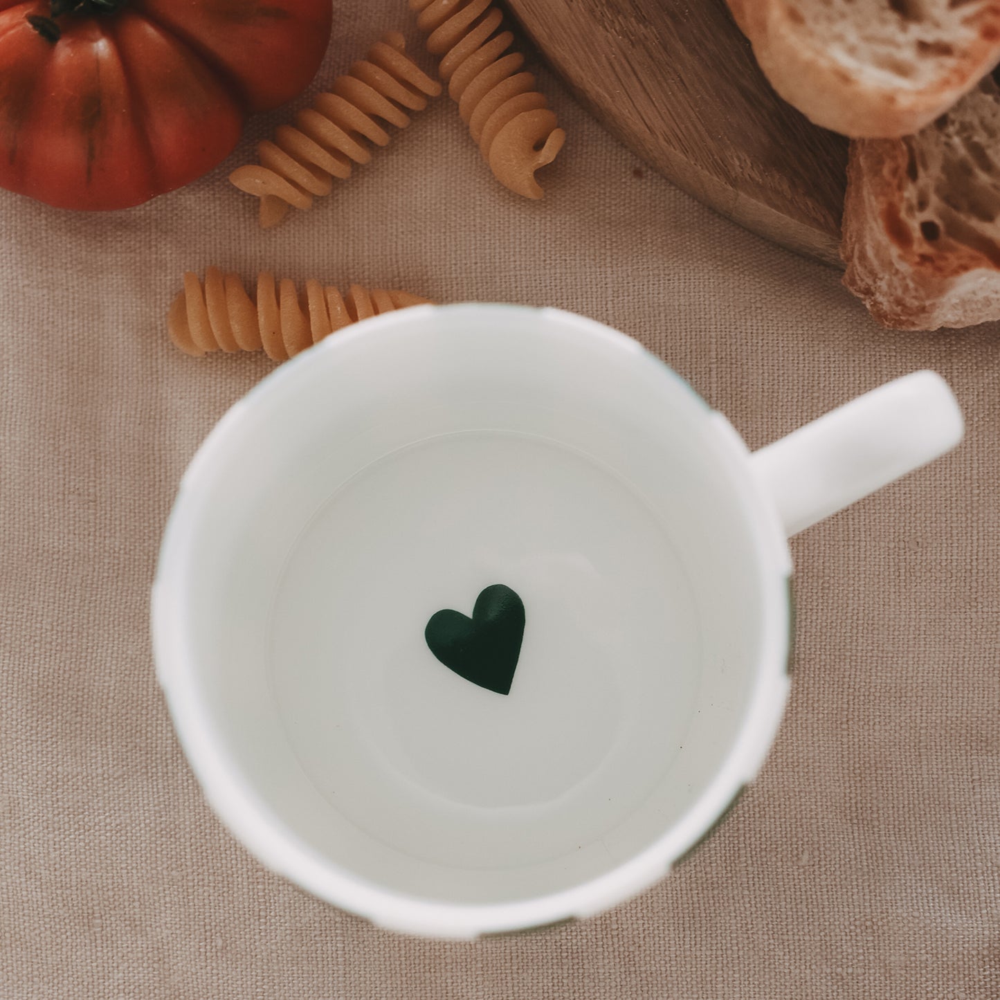 Draufsicht in die kleine weiße Tasse – am Boden ein kleines grünes Herz, umgeben von frischen Zutaten wie Pasta, Tomaten und Brot auf Stoffuntergrund.