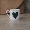leine weiße Tasse mit großem grünem Herz auf der Vorderseite, schlicht inszeniert vor warmem Hintergrund.