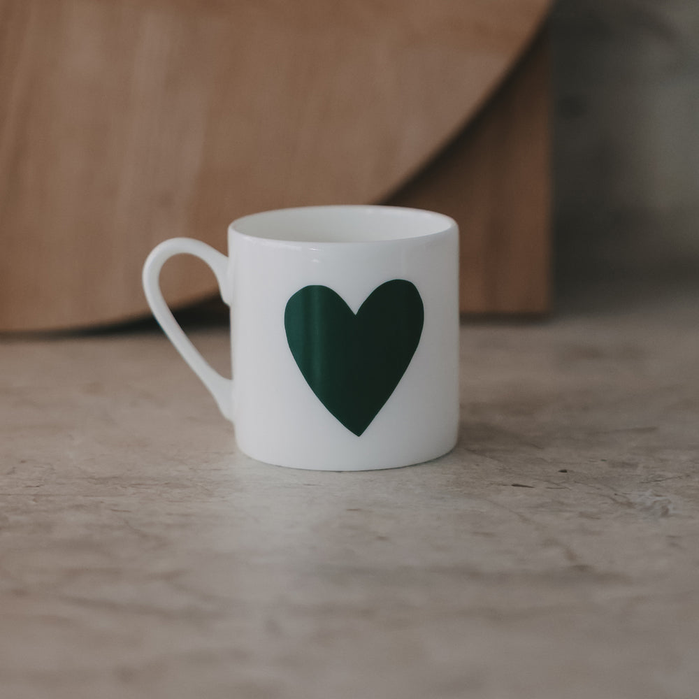 leine weiße Tasse mit großem grünem Herz auf der Vorderseite, schlicht inszeniert vor warmem Hintergrund.