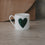 leine weiße Tasse mit großem grünem Herz auf der Vorderseite, schlicht inszeniert vor warmem Hintergrund.