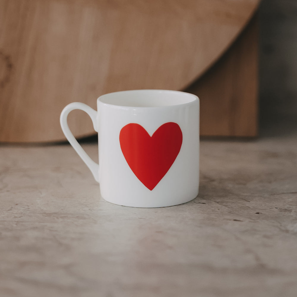 Kleine weiße Tasse mit großem rotem Herz auf der Vorderseite, schlicht inszeniert vor warmem Hintergrund.
