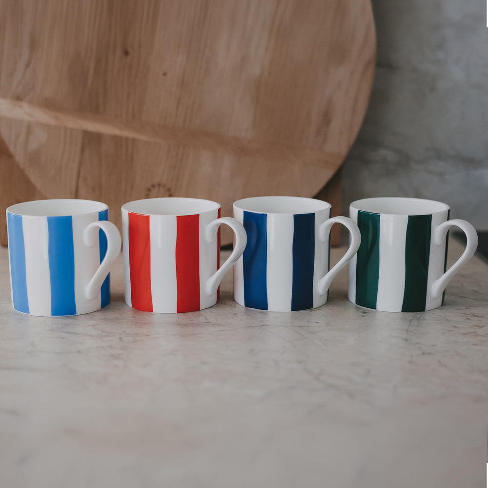 Vier Tassen nebeneinander, jeweils mit breiten Streifen in Blau, Rot, Dunkelblau und Grün auf heller Steinoberfläche.