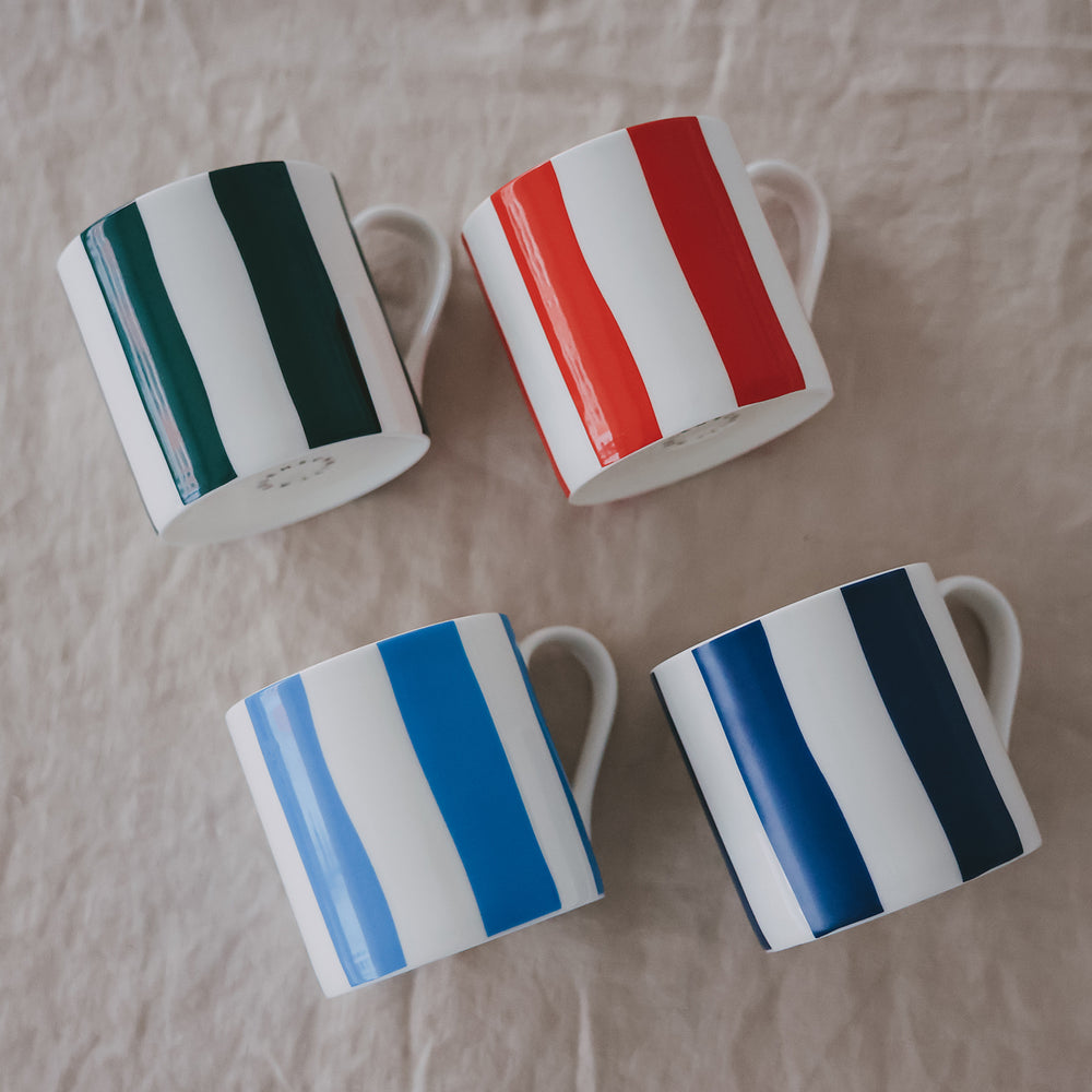 Vier Porzellantassen mit breiten Streifen in Grün, Rot, Hellblau und Dunkelblau, schräg auf hellem Stoff angeordnet.