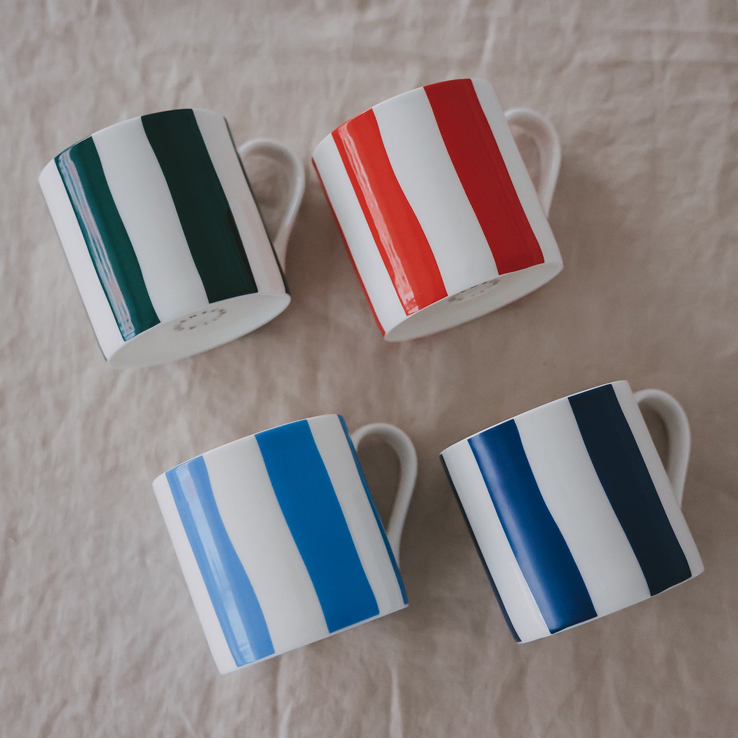 Vier Porzellantassen mit breiten Streifen in Grün, Rot, Hellblau und Dunkelblau, schräg auf hellem Stoff angeordnet.