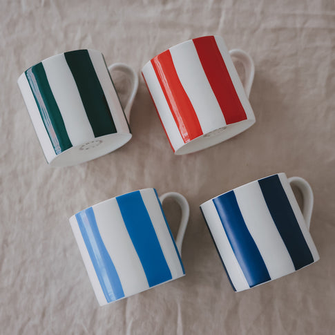 Vier Porzellantassen mit breiten Streifen in Grün, Rot, Hellblau und Dunkelblau, schräg auf hellem Stoff angeordnet.