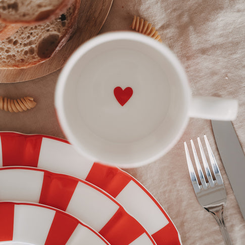 Draufsicht in Tasse mit rotem Herz, umgeben von rot gestreiftem Geschirr und Besteck auf Leinentischdecke