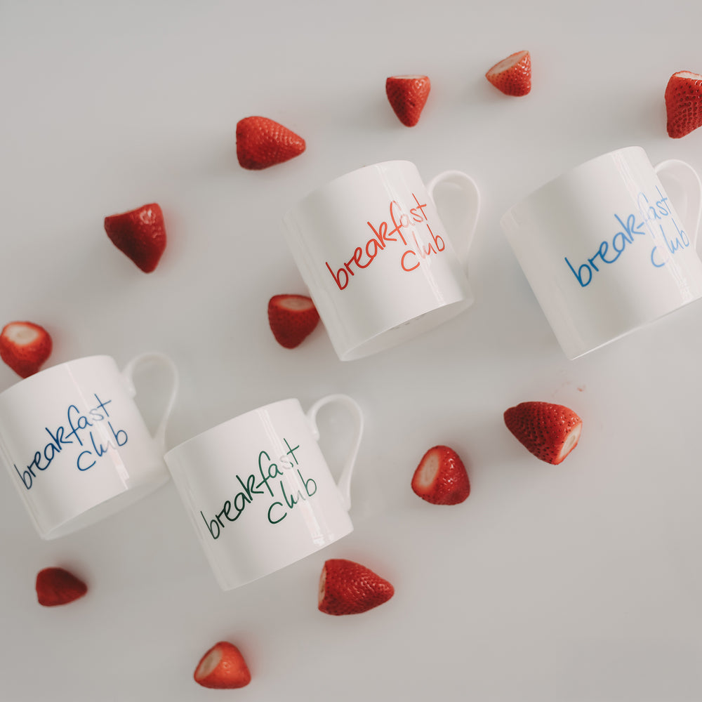 „Breakfast club“-Tassen in Rot, Blau und Grün auf weißem Hintergrund, dekoriert mit halbierten Erdbeeren