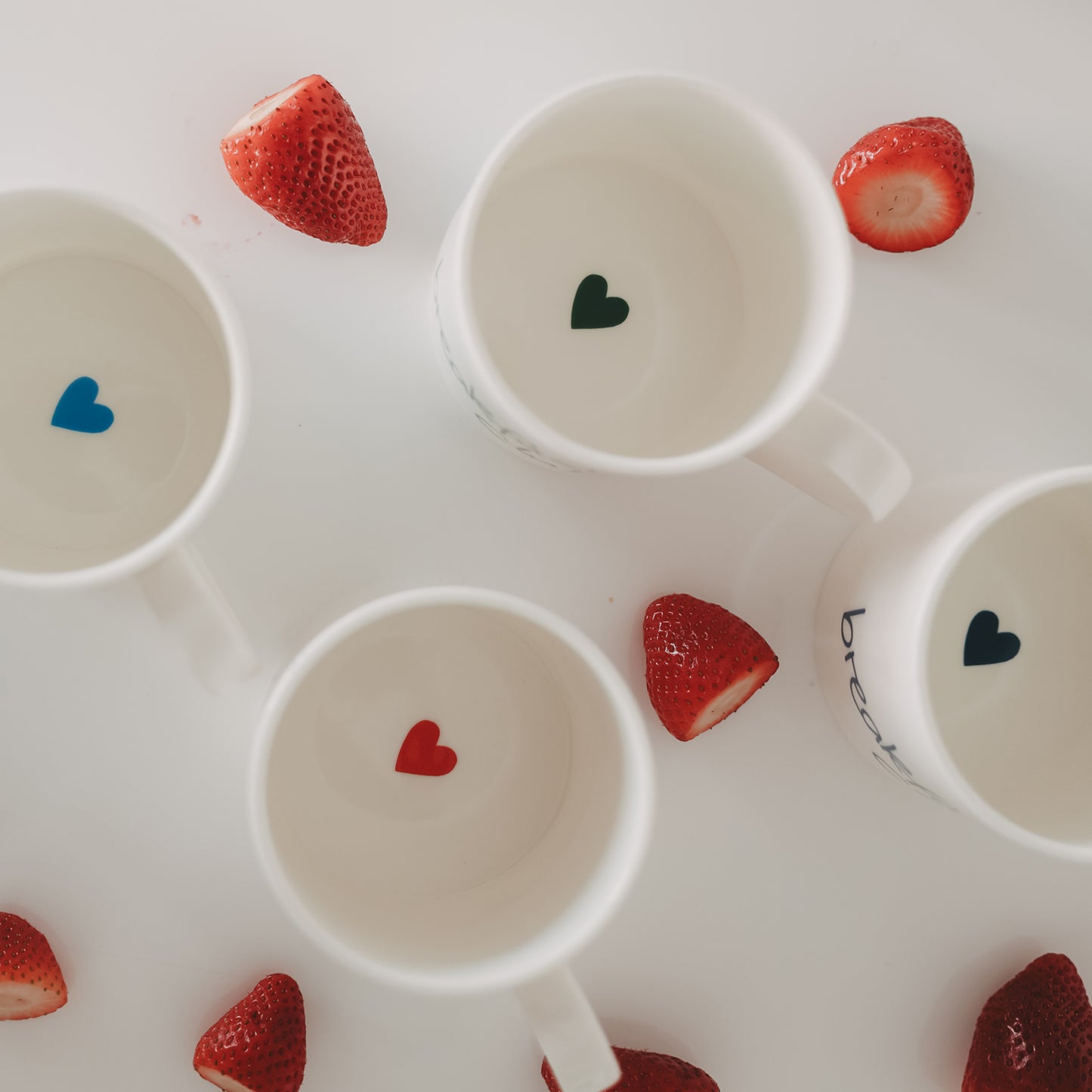 Draufsicht in Tassen mit kleinen Herzmotiven am Boden in Blau, Grün und Rot, umgeben von Erdbeeren