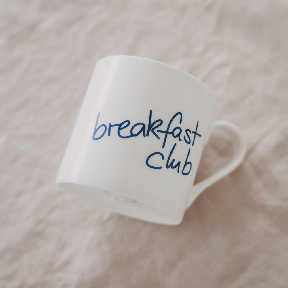 Schräg liegende weiße Tasse mit blauem „breakfast club“-Schriftzug auf hellem Untergrund