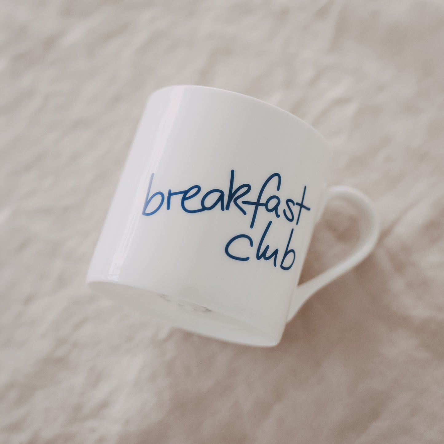 Schräg liegende weiße Tasse mit blauem „breakfast club“-Schriftzug auf hellem Untergrund
