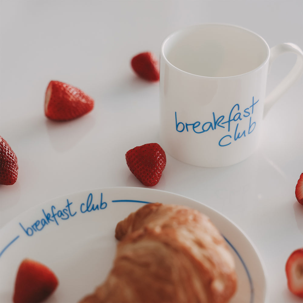 Frühstücksszene mit weißer Tasse mit hellblauem Schriftzug "breakfast club", Teller, Croissant und frischen Erdbeeren