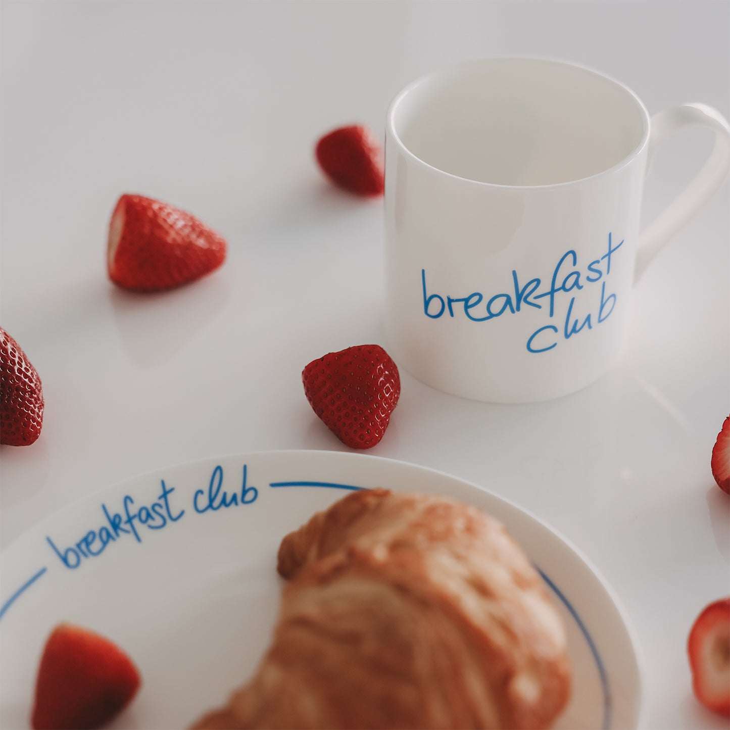 Frühstücksszene mit weißer Tasse mit hellblauem Schriftzug "breakfast club", Teller, Croissant und frischen Erdbeeren