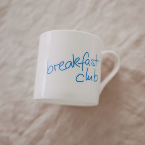 Schräg liegende weiße Tasse mit hellblauem „breakfast club“-Schriftzug auf hellem Leinen-Hintergrund