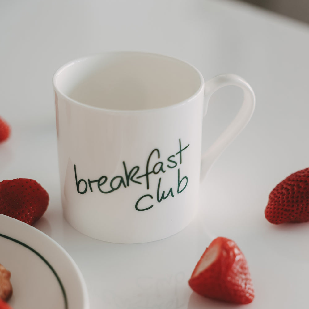 Frühstücksszene mit weißer Tasse mit grünem Schriftzug und Erdbeeren auf hellem Hintergrund