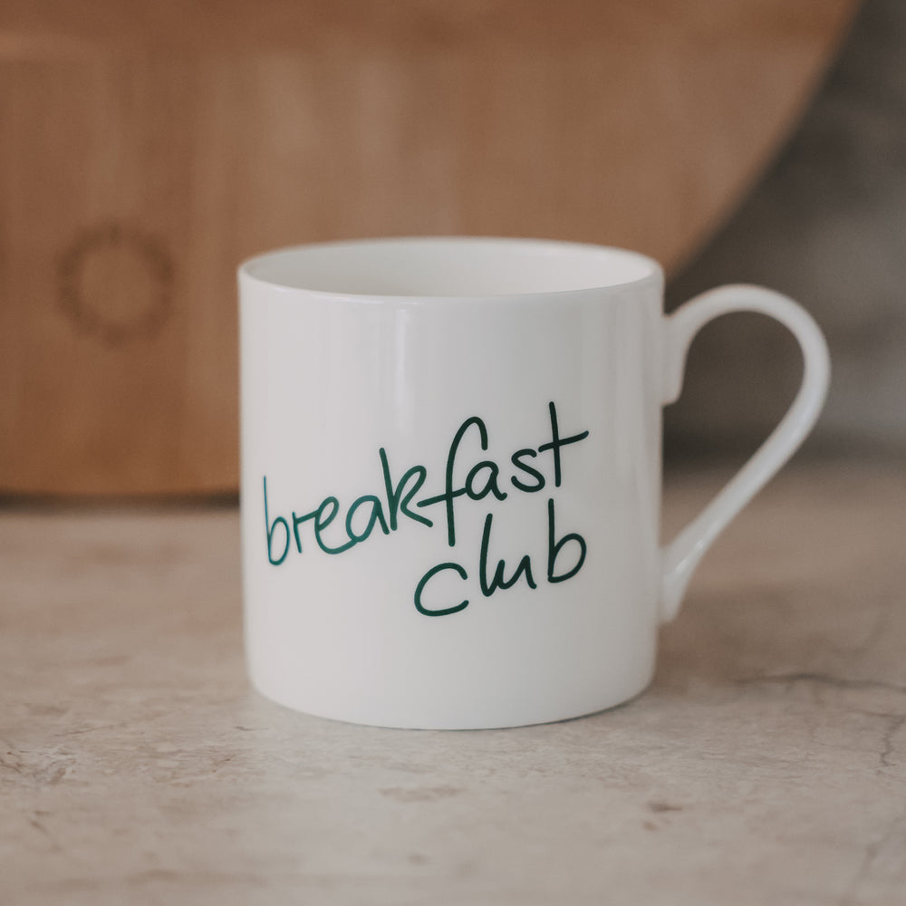 Weiße Tasse mit grünem „breakfast club“-Schriftzug vor warmem Hintergrund