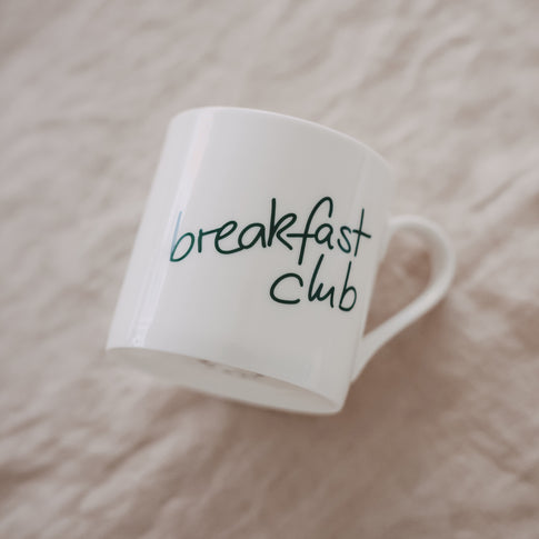 Schräg liegende weiße Tasse mit grünem „breakfast club“-Schriftzug auf hellem Hintergrund