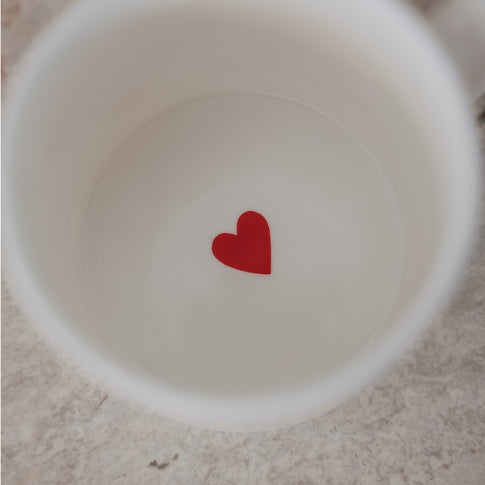 Weiße Tasse mit kleinem rotem Herz im Inneren auf hellem Untergrund