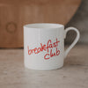 Weiße Tasse mit rotem Schriftzug „breakfast club“ vor warmem Hintergrund