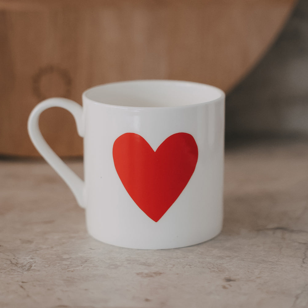 Weiße Tasse mit großem rotem Herz auf der Vorderseite vor warmen Hintergrund